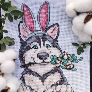 Könnte beinhalten: Kreuzstich-Stickerei eines Husky-Hundes mit rosa Hasenohren, der einen Zweig Grünzeug im Maul hält.
