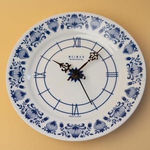 Peut inclure: Une horloge en céramique blanche avec un motif floral bleu sur le bord et des chiffres romains bleus. Les aiguilles de l'horloge sont noires. Le cadran porte l'inscription "WEIMAR" et "MADE IN GDR".