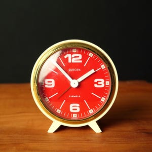 Può includere: Una sveglia Europa vintage con quadrante rosso e numeri bianchi. L'orologio ha una lunetta color oro e una base bianca. Il quadrante mostra il testo "EUROPA" e "2 JEWELS". L'orologio è appoggiato su una superficie di legno.