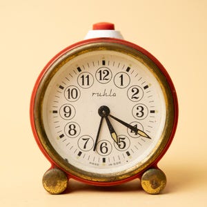 May include: A vintage red and gold alarm clock with a white face and black numerals. The clock face reads "ruhla" and "Made in GDR". The clock has a red top button and two gold-colored feet.