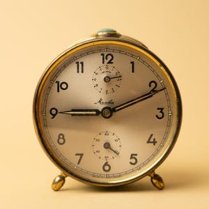Pode incluir: Um despertador dourado vintage com mostrador creme. O relógio apresenta algarismos pretos, ponteiros e a marca "Mantha". Possui dois mostradores menores e repousa sobre dois pequenos pés dourados. A hora é aproximadamente 8h46.