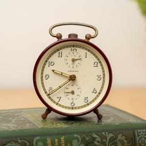 Può includere: Una sveglia vintage con quadrante color crema, numeri neri e lancette dorate. L'orologio ha una cornice bordeaux e una maniglia dorata. È appoggiato su un libro verde con scritte dorate. L'orologio misura circa 10 cm di altezza.