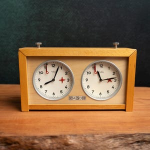 Può includere: Un orologio da scacchi vintage con due quadranti circolari. L'orologio ha una cornice in legno chiaro e una base in legno chiaro. I quadranti hanno lancette nere, dettagli rossi e la parola "GARDE" è stampata in basso.