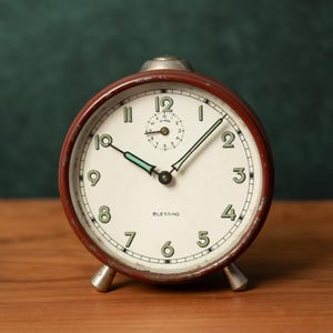 Pode incluir: Um despertador vintage vermelho com um mostrador branco e ponteiros pretos. O mostrador do relógio tem números pretos e a palavra "Blessing" impressa nele.