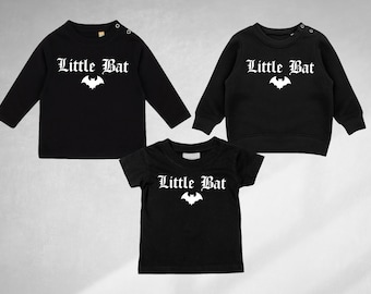 Może przedstawiać: Trzy czarne koszulki z napisem "Little Bat" w białym gotyckim foncie i białym nadrukiem nietoperza. Koszulki są z długim rękawem, krótkim rękawem i bluza.