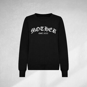 K&ouml;nnte beinhalten: Schwarzer Pullover mit einem wei&szlig;en Grafikdruck, der "MOTHER EST 2022" in einer gotischen Schriftart zeigt.