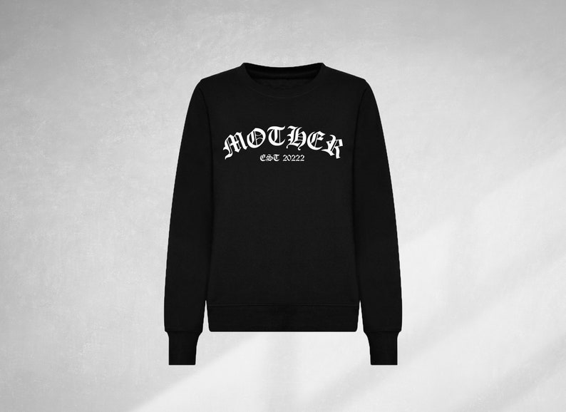 K&ouml;nnte beinhalten: Schwarzer Pullover mit einem wei&szlig;en Grafikdruck, der "MOTHER EST 2022" in einer gotischen Schriftart zeigt.