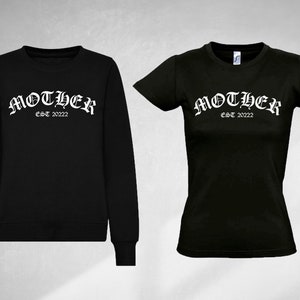K&ouml;nnte beinhalten: Schwarzer Pullover und T-Shirt mit dem Text "MOTHER EST 2022" in wei&szlig;en gotischen Buchstaben.
