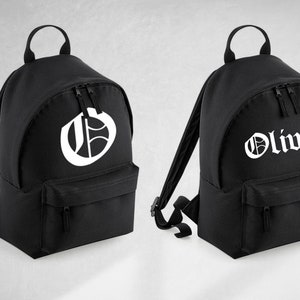 May include: Two black canvas backpacks with white lettering. One backpack has a large letter "G" on the front. The other backpack has the name "Oliver" in a gothic font.
