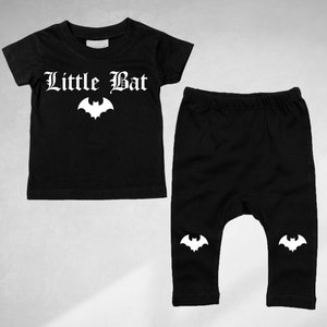 Peut inclure: Ensemble bébé noir avec un motif de chauve-souris blanc et le texte "Little Bat" sur le t-shirt. Le pantalon a un motif de chauve-souris blanc sur chaque jambe.