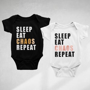 以下が含まれることがあります： 黒と白のベビーロンパース2枚。それぞれに「SLEEP EAT CHAOS REPEAT」の文字がプリントされています。黒いロンパースには「CHAOS」の文字が金色で、白いロンパースにはピンク色でプリントされています。