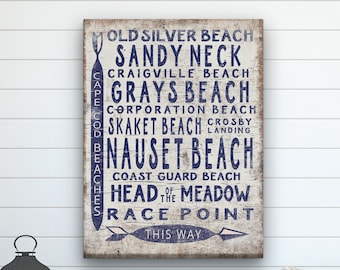 May include: A white wooden sign with blue text listing various beach names, including Old Silver Beach, Sandy Neck, Grays Beach, Nauset Beach, and Race Point. The sign also includes the text "This Way" with an arrow pointing to the right.