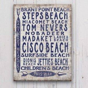 May include: A white wooden sign with blue text listing the names of beaches on Nantucket Island, Massachusetts. The sign includes the text "BRANT POINT BEACH", "STEPS BEACH", "MIACOMET BEACH", "TOM NEVERS", "NOBADEER", "MADAKET LADIES BEACH", "CISCO BEACH", "SURFSIDE BEACH", "DIONIS JETTIES BEACH", "CHILDREN'S BEACH", and "THIS WAY".