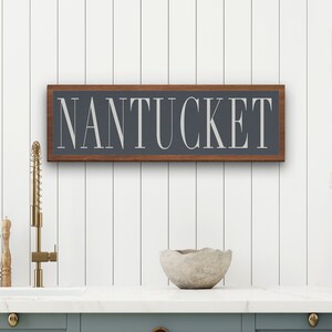 May include: A wooden sign with a dark gray background and a white inscription that reads "NANTUCKET". The sign is framed in a dark wood frame and is hanging on a white wall.