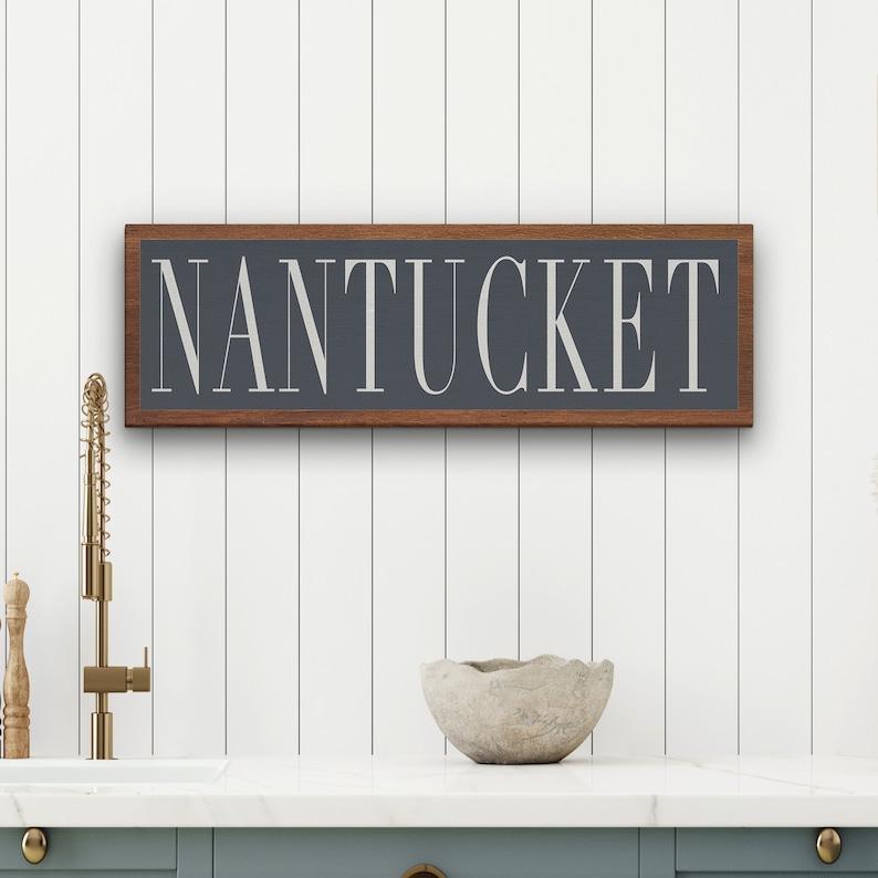 May include: A wooden sign with a dark gray background and a white inscription that reads "NANTUCKET". The sign is framed in a dark wood frame and is hanging on a white wall.