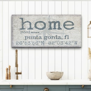 May include: A rustic wooden sign with the word "home" in blue text, a compass rose, and the text "punta gorda, fl 26°53'56"N 82°03'42"W".