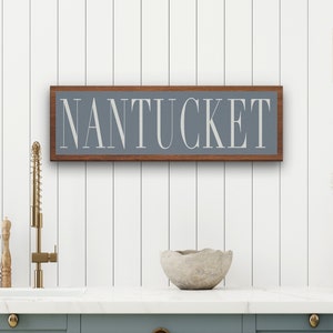 May include: A wooden sign with a gray background and the word "NANTUCKET" in white lettering. The sign is framed in a dark wood frame.