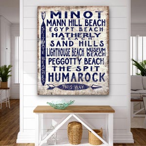 May include: A rectangular wooden sign with a distressed finish, featuring navy blue text. The sign lists various beach names, including "Minot" and "Lighthouse Beach." A directional arrow points towards the bottom. The sign is mounted on a white wall.