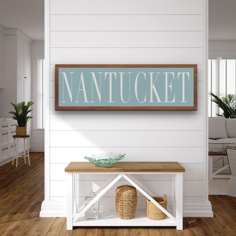 May include: A wooden sign with a light blue background and white text that reads "NANTUCKET". The sign is hanging on a white wall with white shiplap siding. The sign is above a white console table with a wooden top. The table has two shelves and is decorated with a glass bowl, a wicker basket, and a white lantern.