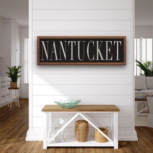 May include: A wooden sign with a black background and white lettering that reads "NANTUCKET". The sign is hanging on a white shiplap wall above a wooden console table.
