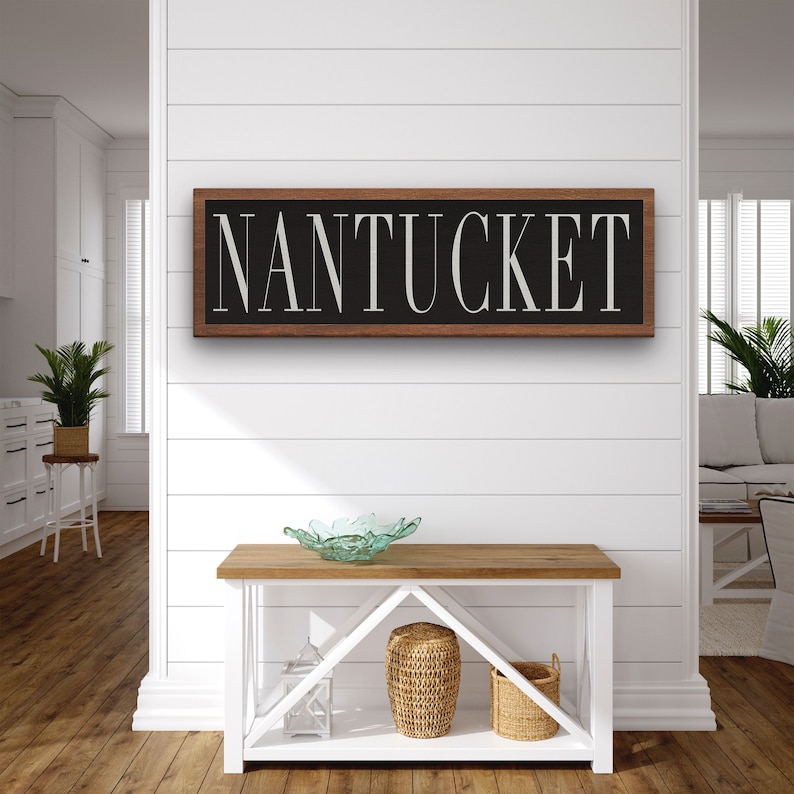May include: A wooden sign with a black background and white lettering that reads "NANTUCKET". The sign is hanging on a white shiplap wall above a wooden console table.