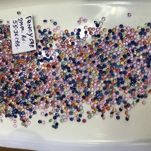 Könnte beinhalten: Eine Sammlung loser, runder, facettierter Edelsteine in verschiedenen Blautönen, Rosa, Gelb und Orange. Die Edelsteine haben einen Durchmesser von etwa 2 mm und sind mit "Fancy Sap. 2mm - Rd. 5.5-3.6 ctrs." beschriftet.