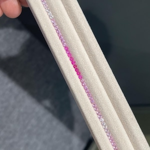 May include: A close-up of a row of pink sapphires of varying shades, set in a white fabric display.