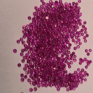 May include: A scattering of small, round, faceted gemstones in a vibrant magenta colour. The gemstones are spread across a white surface, creating a sparkling display. These could be rubies or other precious stones.