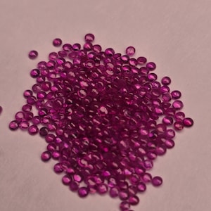 May include: A pile of small, round, faceted, deep pink gemstones. The gems are scattered across a light pink surface, with a slight variation in color and shine. The gemstones appear to be polished and ready for use in jewelry or other crafts.