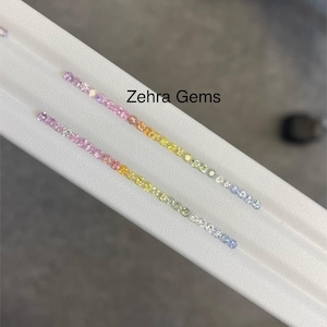 May include: Two rows of colorful gemstones arranged in a gradient pattern, from pink to yellow and blue. The gemstones are displayed on a white surface, with the text "Zehra Gems" visible.
