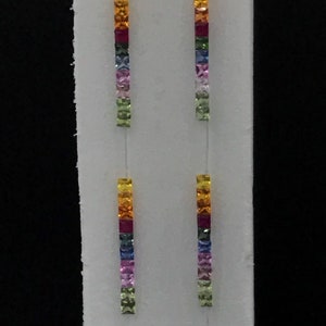 Op de afbeelding: Twee paar oorbellen met een regenboog van vierkante edelstenen. Elke oorbel heeft een rij gele, oranje, rode, paarse, blauwe, groene en lichtgroene edelstenen.