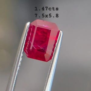May include: A vibrant red gemstone, emerald cut, held by tweezers. The gem is 7.5mm x 5.8mm and weighs 1.47 carats. The stone has a deep, saturated color and is displayed against a blurred background.