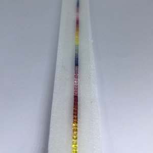 May include: A row of small, square-shaped gemstones in various colors, including yellow, orange, red, pink, purple, and blue, arranged in a vertical line on a white background.