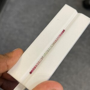 May include: A row of small, round gemstones in a gradient of pink, white, and green colors. The gemstones are set in a thin metal band.