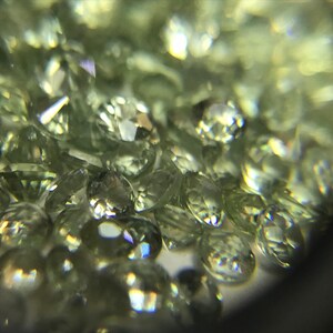 May include: A close-up view of a group of small, round, light green gemstones. The gemstones are faceted and have a shiny, reflective surface.