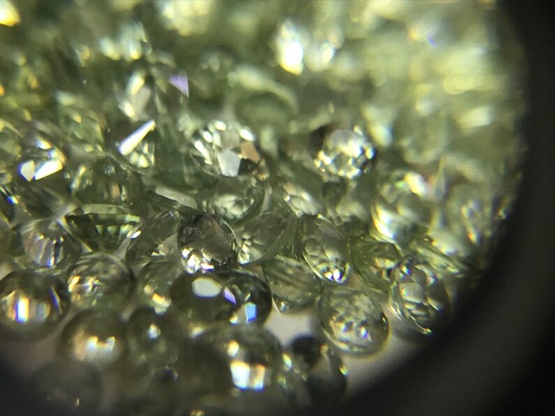 May include: A close-up view of a group of small, round, light green gemstones. The gemstones are faceted and have a shiny, reflective surface.