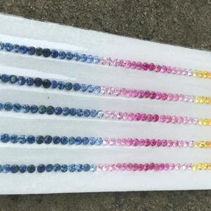 May include: Rows of small, round gemstones in a variety of colors, including green, blue, pink, yellow, and orange. The gemstones are arranged in a gradient pattern on a white surface, creating a visually appealing display.