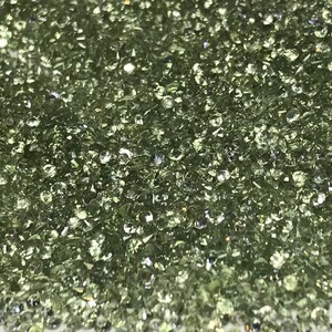 May include: A close-up view of a large quantity of small, round, light green gemstones. The gemstones are scattered across a white surface.