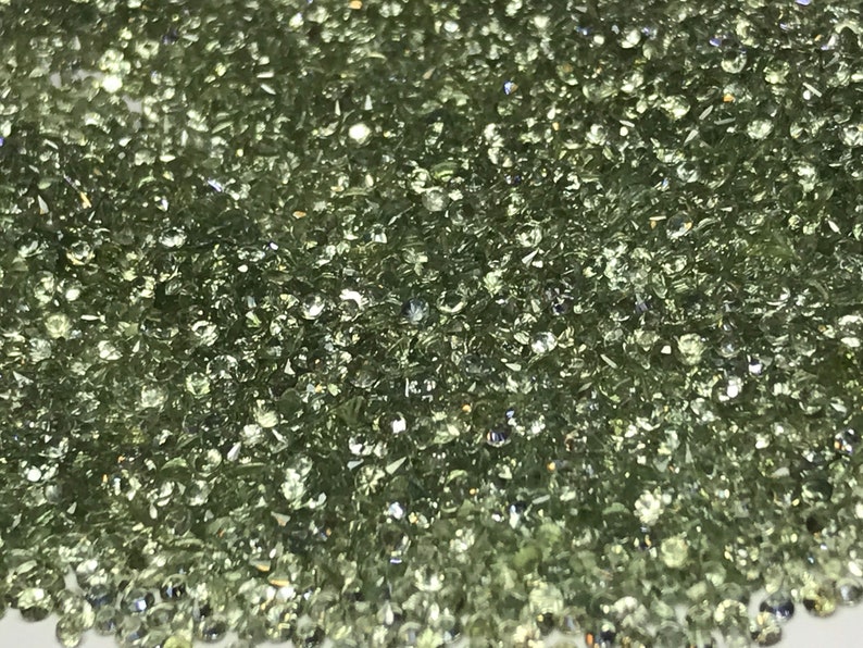 May include: A close-up view of a large quantity of small, round, light green gemstones. The gemstones are scattered across a white surface.