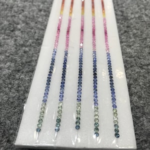 Könnte beinhalten: Fünf Stränge abgestufter Saphirperlen in Grün-, Blau-, Rosa- und Orangetönen. Die Perlen sind in Reihen auf einem weißen Schaumstoffuntergrund angeordnet.