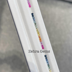 May include: Two rows of small, round gemstones arranged in a rainbow color gradient. The gemstones are on a white surface with the text "Zehra Gems" printed below.