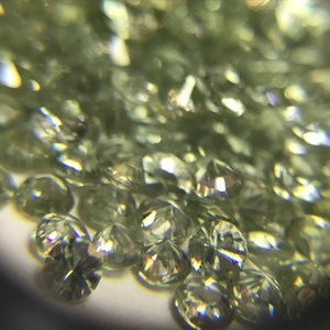 May include: A close-up view of a pile of small, round, green gemstones. The gemstones are faceted and have a shiny, reflective surface.