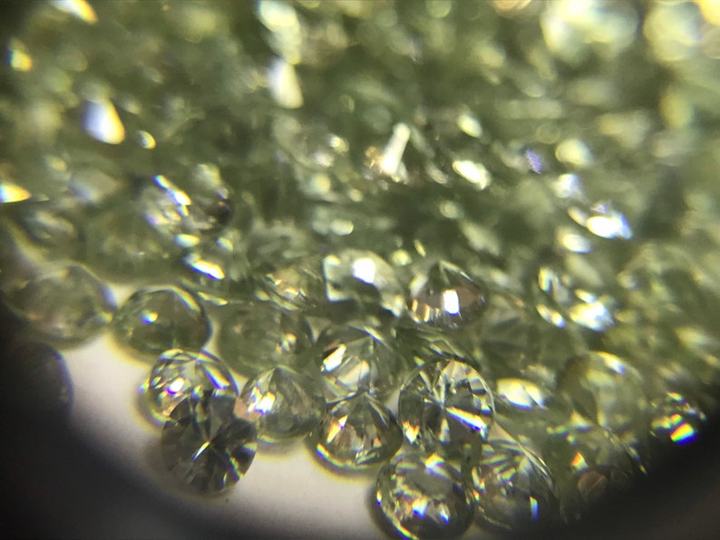 May include: A close-up view of a pile of small, round, green gemstones. The gemstones are faceted and have a shiny, reflective surface.