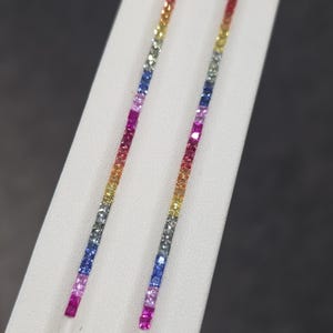May include: A pair of long, linear drop earrings featuring a vertical row of square-cut gemstones in a vibrant rainbow gradient, transitioning through shades of red, orange, yellow, green, blue, and purple in a polished metal setting.