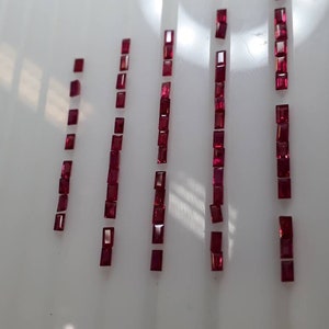 May include: A collection of 10 rows of small, rectangular, red gemstones. The gemstones are arranged in vertical rows on a white surface.
