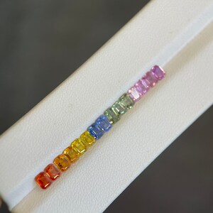 May include: A row of colorful gemstones arranged in a rainbow spectrum. The gems transition from red to orange, yellow, green, blue, and purple. Each gem is rectangular and set on a white surface, showcasing their vibrant hues. The gemstones are likely sapphires.