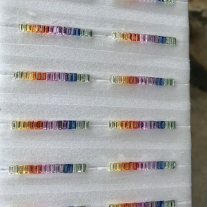 Puede incluir: Una tabla de espuma blanca con filas de pequeñas piedras preciosas rectangulares multicolores. Las piedras preciosas están dispuestas en orden de arcoíris, de amarillo a azul.