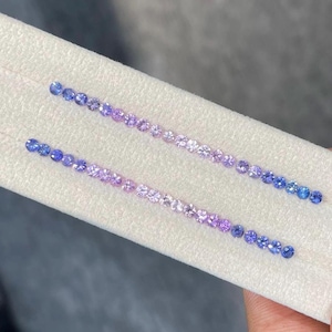 Puede incluir: Dos filas de piedras preciosas redondas y facetadas dispuestas en un degradado de azul oscuro a morado claro. Las piedras preciosas se muestran sobre una superficie blanca.