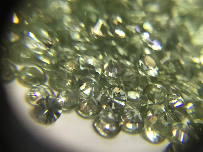May include: A close-up view of a pile of small, round, faceted green gemstones. The gemstones are translucent and have a slight shimmer.