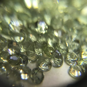 May include: A close-up view of a pile of small, faceted, light green gemstones. The gemstones are oval-shaped and have a shiny, reflective surface.